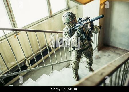 warlike soldier man goes up the stair looking for enemy or danger, holding weapon in hands, always ready to shoot Stock Photo