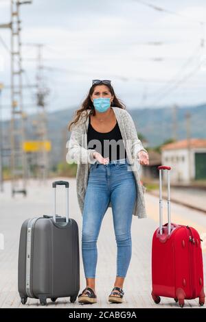 Young woman with a surgical mask surrounded by two suitcases makes a questioning gesture at the camera. Travel and safety concept. Stock Photo