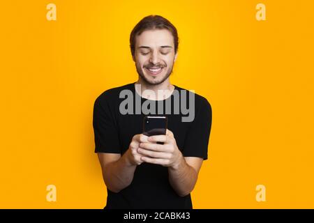 Bearded man with long hair is chatting on mobile in black clothes and smile on a yellow studio wall Stock Photo