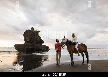 Asia, Indonesia, Bali, young Caucasian man with a beautiful young caucasian woman sitting on a horse, on a beach at sunset, with typical balinese rock Stock Photo