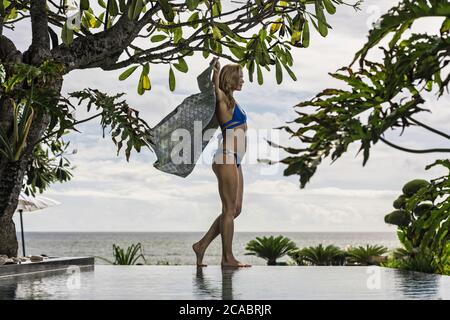 Asia, Indonesia, Bali, young beautiful Caucasian woman, wearing blue bikini, walking along on edge of infinity pool, holding a sarong, with tropical p Stock Photo