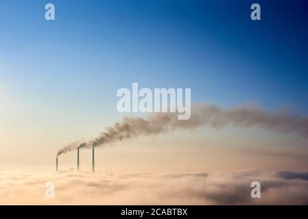 Tops of three smoking stacks of thermal power station on the horizon taken from the hill, pipes in morning fog on blue sky, concept of dangerous emission in atmosphere, energy generation, ecology Stock Photo