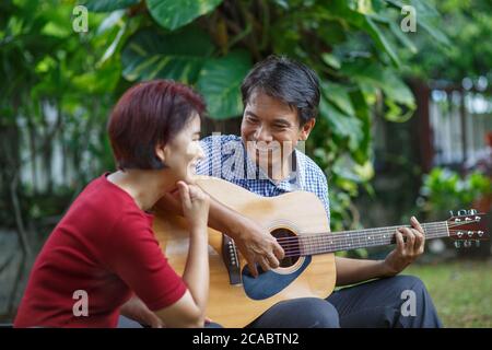 Middle aged  couple playing guitar while relax sitting on bench in backyard. Stock Photo