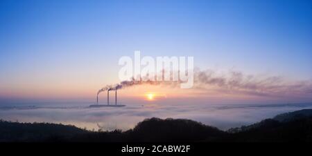 Panoramic view of three smoking stacks of thermal power station on the horizon taken from the hill, pipes are in evening fog on blue sky, copy space. Concept of ecology and environmental pollution. Stock Photo