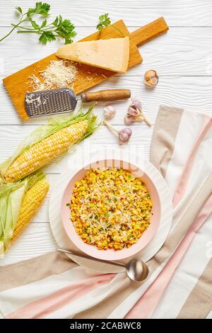 Parmesan Cilantro Corn in a pink bowl on a white wooden table with ingredients, vertical view from above, close-up Stock Photo