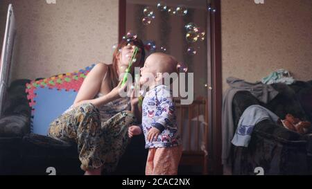 a young beautiful woman blowing soap bubbles and her little baby boy plays catching them happily smiling inside room in slow motion 4K video Stock Photo