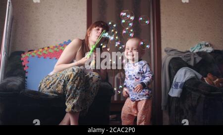 little baby boy playing with his mother who blowing soap bubbles while kid happily smiling catching them inside room in slow motion 4K video Stock Photo