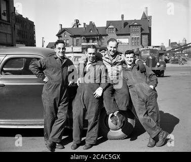1940s, historical, outside in the square of an midwest industrial town, four motor mechanics in their overalls leaning against a motorcar pose for a photograph, USA. A sign for the Prens Carting Co can be seen in the background. A Socony badge can be seen on one of the mechanic's overalls, the shortened name for the 'Standard Oil Co of New York'.