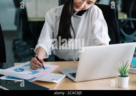 Young accountant hand holding smartphone to call marketing consultants and using laptop computer to analyze sales growth in the global workplace Stock Photo