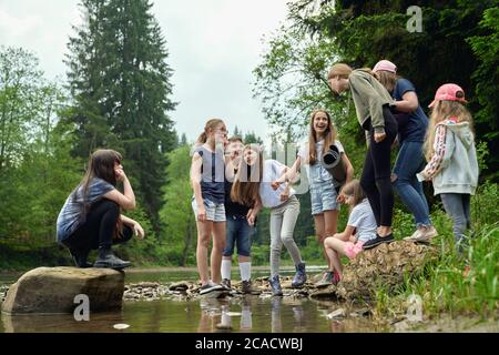 From below view of laughing teens in casual suits standing on rocks in river water in summer day. Young friends having fun outdoors near green forest. Concept of friendship, nature, camping. Stock Photo