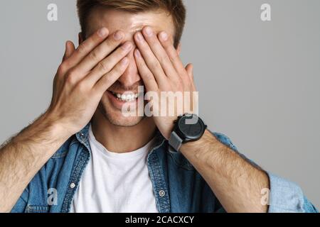 Portrait of cheerful unshaven man laughing and covering his eyes isolated over grey background Stock Photo