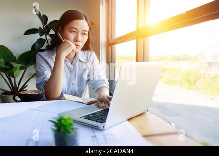 Serious Asian female architects working under pressure using laptop for researching the architectural analyze project sit near the window at home Stock Photo