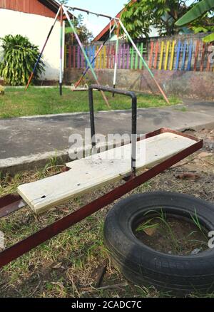 broken board of the seesaw with broken tire for safety pads in an empty playground without children Stock Photo