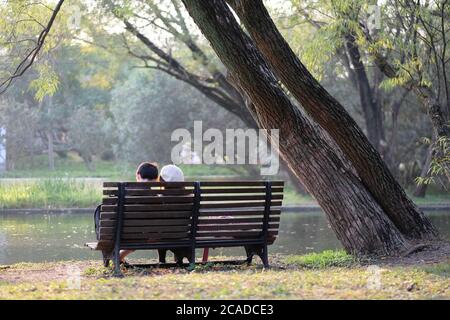 a pair of lovers sitting on wooden bench in park under sunshine. Lake water and trees around Stock Photo