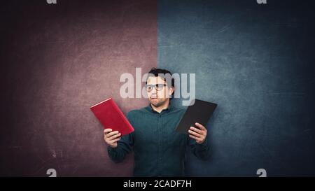 Pensive male student chooses between a traditional book and electronic PC tablet over a split wall with red and blue sides. Future education concept. Stock Photo
