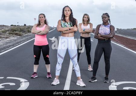 Full body group of confident multiracial sportswomen with crossed arms looking at camera while standing on asphalt road during running session Stock Photo