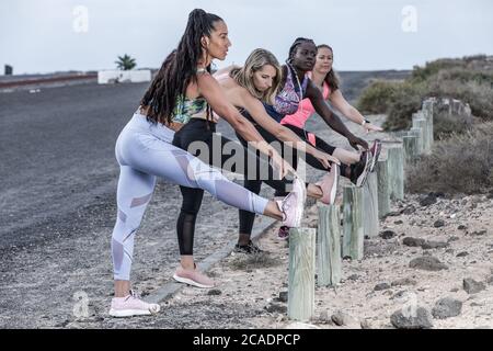 Side view of full body multiethnic female friends in sportswear keeping legs on poles and bending forward while warming up before running session toge Stock Photo