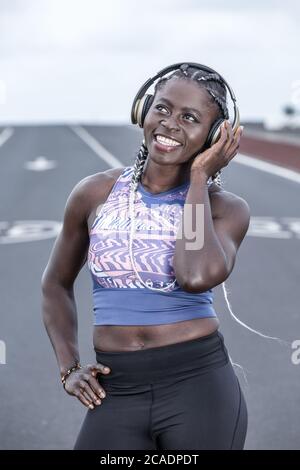 Positive African American female smiling and looking up while listening to favorite music in headphones during break in outdoor workout Stock Photo