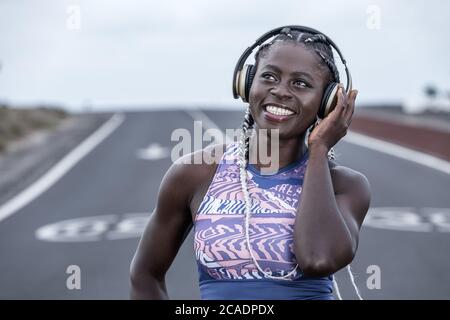 Optimistic African American female athlete smiling and looking away while listening to music in earphones during break in outdoors training on blurred Stock Photo