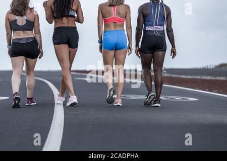 Back view of crop diverse female athletes in sportswear walking on asphalt road during outdoor workout in outskirts Stock Photo