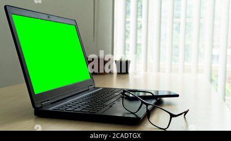 laptop on wooden desk with green screen glasses and smartphone with blur background Stock Photo