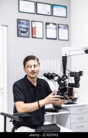 Mature stomatologist posing on workplace, near dental microscope, leaning on dentist chair. Handsome, bearded stomatologist wearing in white medical c Stock Photo