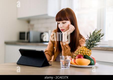 Horizontal shot of cute dark haired Caucasian businesswoman wearing domestic casual wear, sitting at table in kitchen and using digital tablet ipad Stock Photo