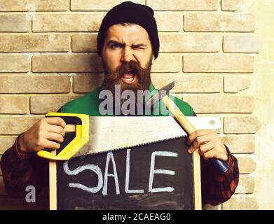 bearded worker man, long beard, brutal caucasian hipster with moustache, black cap, holding various building tools: saw, hammer, board with inscription sale, satisfied face, brick wall background Stock Photo