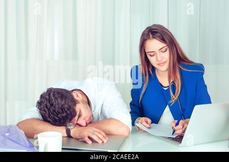 Bored lazy man sleeping at table with smart creative woman working sitting near at table in office living room at home. Overworking late hours working Stock Photo