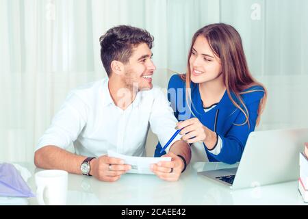 Young smiling man and woman looking at each other while working on paper in team. Happy couple colleagues sharing work sitting at table in office livi Stock Photo