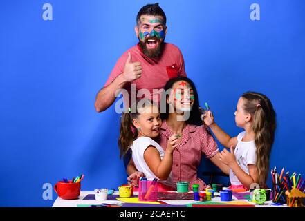 Parents and children painted with gouache on blue background. Girls, man and woman with cheerful faces by desk with paints. Artists family paints ladys face with their fingers. Art and craft concept Stock Photo