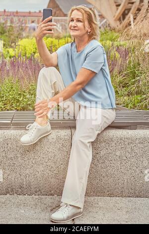 Elderly white attractive lady about 60 years old is sitting on the bench in the public park and making photos by her cell phone. She wears elegant fas Stock Photo