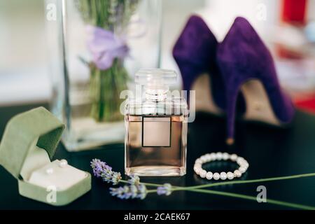 A bottle of perfume on the table with lilac women's high-heeled shoes, a bracelet made of white beads, a box with stud earrings and a vase of flowers. Stock Photo