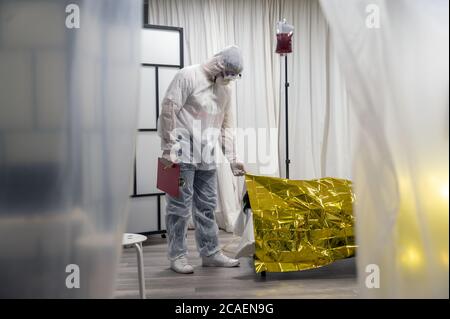 Alert pandemic Covid-19. Doctor in protective white suit, glasses and mask covers a lifeless body for Corona Virus epidemy. Stock Photo