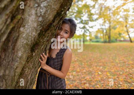 Young happy Asian woman smiling while playfully hiding behind tree Stock Photo