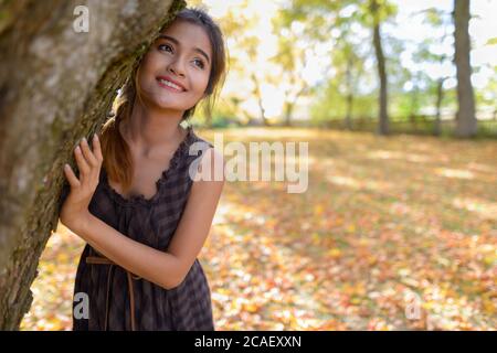 Young happy Asian woman smiling while looking up playfully hiding behind tree Stock Photo