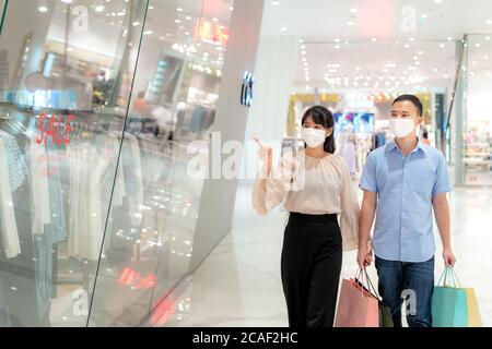 Asian couple wearing mask over their face looking new collection dress at shopping mall with shopping bag for healthcare and prevention from coronavir Stock Photo