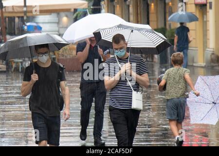 Belgrade, Serbia - August 5, 2020: Two young men wearing face surgical masks and eyeglasses walking under umbrellas in the crowd at the pedestrian str Stock Photo