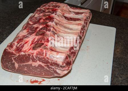 Close up of a large raw beef prime rib roast sitting on a white plastic cutting board in a restaurant kitchen. The meat is marbled with six ribs. Stock Photo