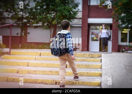 Back view child boy with bag goes to elementary school and masked woman comes out of the door. Stock Photo