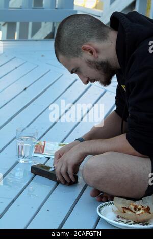 Young man sharpening a knife using a whetstone while sitting in a wood deck Stock Photo