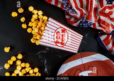 Yellow popcorn in striped boxes spilled on black background with american football plate. Watching sport game concept. Stock Photo