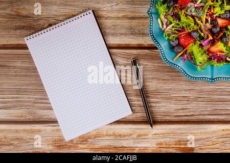 Notepad and pen with fruit and vegetable salad on wooden background. Stock Photo