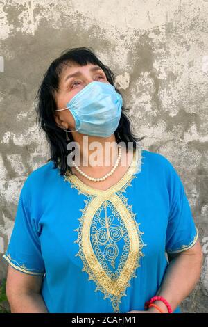 Asian or caucasian ethnicity mature woman in blue dress with medical mask on his face to protect COVID-19, looking up against background of gray plast Stock Photo