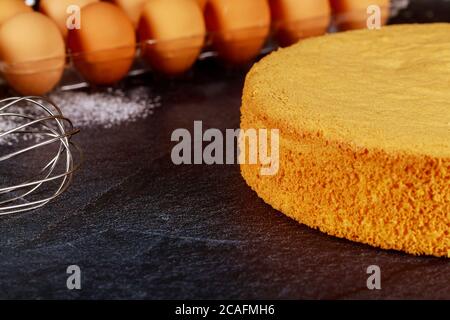 Delicious sponge cake on dark table with raw eggs and whisk. Bakery concept. Stock Photo