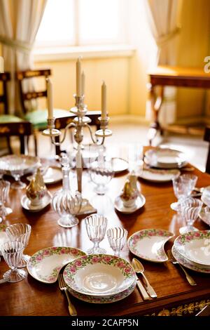 Festive table setting with clean empty plates, spoons, knives, wine glasses, decanters and a candlestick in the center on a wooden table in the room. Stock Photo