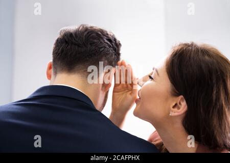 Woman Whispering Gossip In Friend's Ear At Workplace In Office Stock Photo