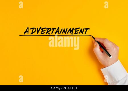 Male hand is writing the word advertainment on yellow background. Business buzzword concept that intagrates advertising and entertainment. Stock Photo