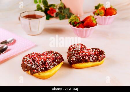 Two heart shaped donuts with white glaze and pink, red sprinkles on a platet with strawberry and cup of tea. Valentine Day concept. Stock Photo
