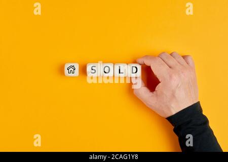 Concept of selling or buying a house or real estate. House icon and sold word on wooden cubes with a male hand placing the last letter. Stock Photo
